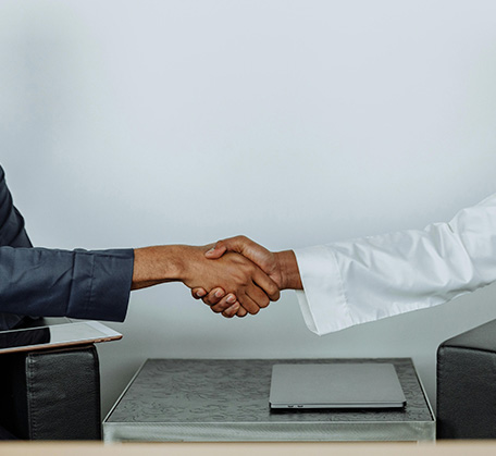 Two people shaking hands across a small table with a laptop, one in a suit and the other in white attire.