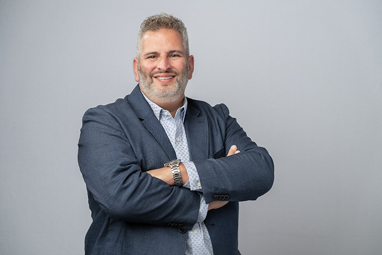 Chris Kourtakis smiling with arms crossed, wearing a blazer and patterned shirt against a light background.