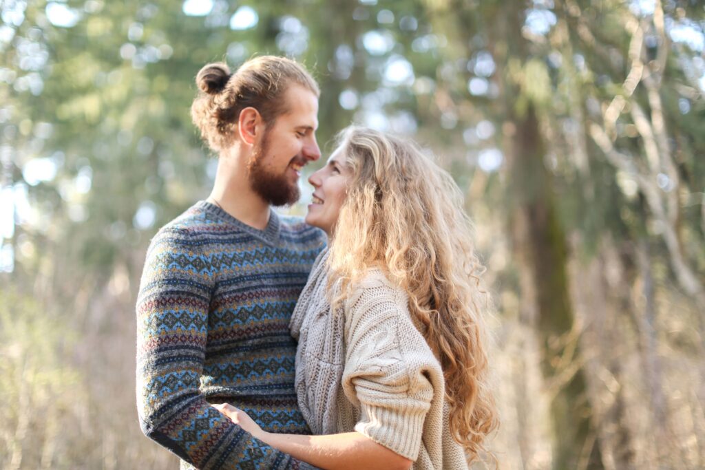 Couple in knitwear sharing affectionate gaze amidst woodland backdrop.