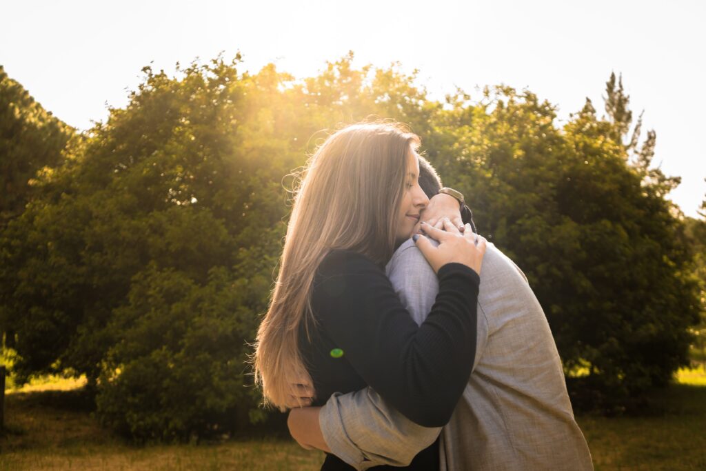 A couple shares a warm embrace under the bright sun in a peaceful park setting.