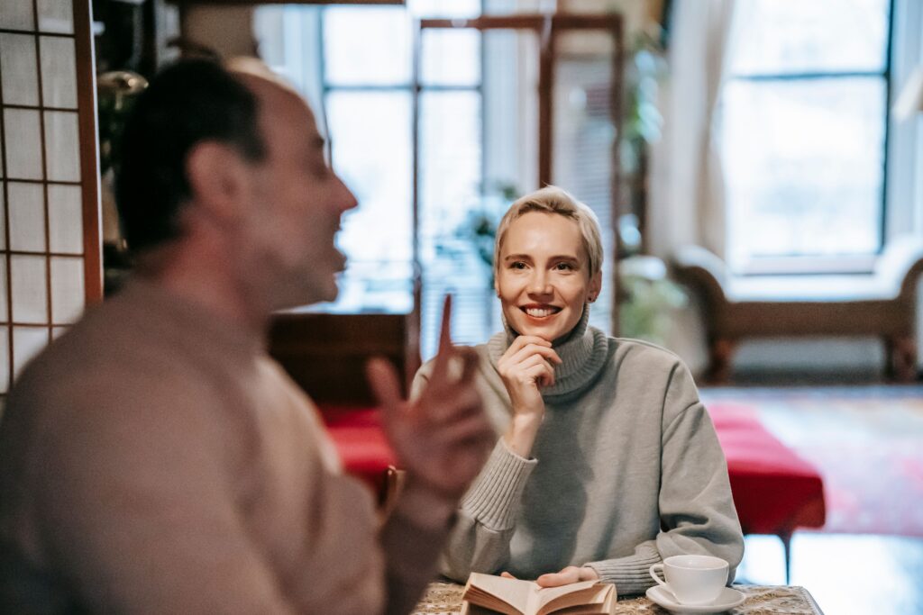 Optimistic adult lady sitting at table with book and touching chin while communicating with unrecognizable husband during weekend at home